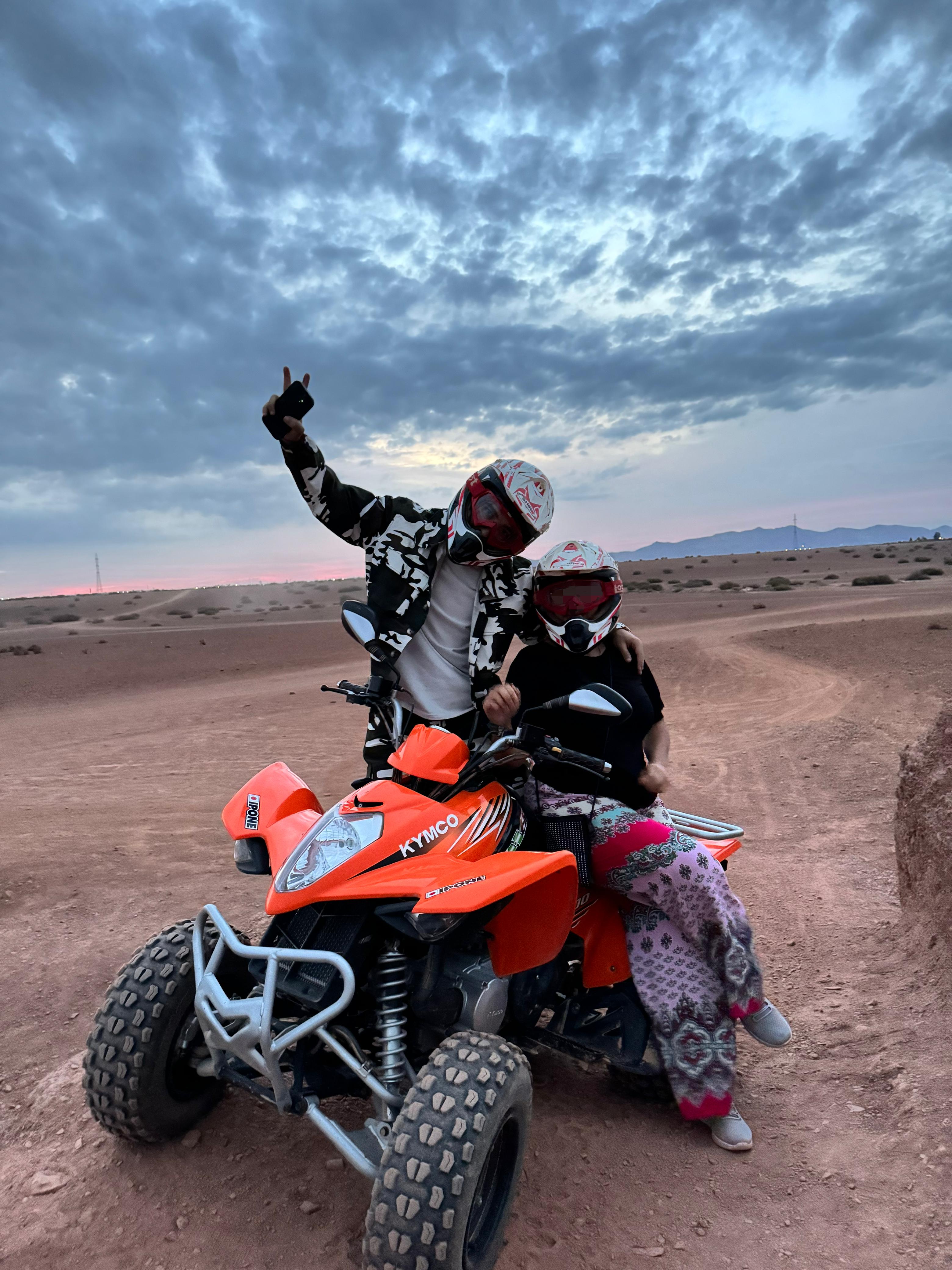 Two people on red ATV making peace signs under dramatic cloudy sky at sunset